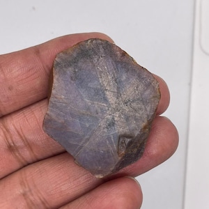 Pode incluir: Uma pedra preciosa de safira estrela bruta, plana e de formato hexagonal, com um tom translúcido de roxo empoeirado e cinza. A superfície exibe um padrão natural distinto de estrela de seis pontas que irradia do centro, com bordas ásperas e tons terrosos.
