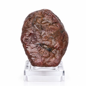 Peut inclure: Une pierre brute brun-rouge foncé avec une surface texturée et irrégulière présentant des fissures naturelles, montée sur un socle carré en acrylique transparent. La roche affiche des tons marron et bordeaux avec des variations minérales.