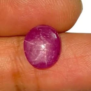 Può includere: Una gemma di rubino cabochon di forma ovale caratterizzata da un distinto effetto a stella a sei raggi sulla sua superficie rosso magenta intenso. La pietra presenta una cupola liscia e lucida con inclusioni interne visibili e lucentezza.