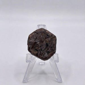 以下が含まれることがあります： 透明なアクリル製の三脚スタンドに飾られた、暗い茶色でざらざらした質感の六角形の石の標本。石の表面は不均一でくぼみがあり、自然な色合いと深みの変化が見られ、透明なサポートの上にしっかりと置かれています。