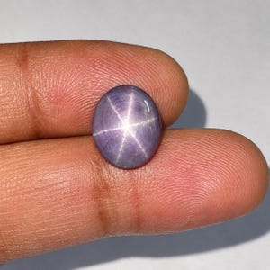 Può includere: Una gemma a cabochon di forma ovale liscia con una tonalità viola lavanda traslucida. La pietra presenta un distinto motivo a stella a sei raggi, noto come asterismo, centrato sulla sua superficie lucida, creando un effetto luminoso.