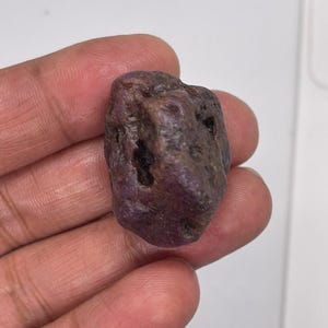 Puede incluir: Una piedra preciosa de rubí en bruto y sin pulir con un tono rojo violáceo intenso. La piedra presenta una forma orgánica irregular con una superficie texturizada, que incluye varias pequeñas hendiduras y hoyos naturales oscuros.