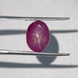 Pode incluir: Uma gema de rubi cabochão de formato oval com um efeito de estrela de seis raios distinto visível na sua superfície. A pedra vermelho-rosada profunda é mantida firmemente entre as pontas metálicas de uma pinça prateada, exibindo a sua cúpula polida.