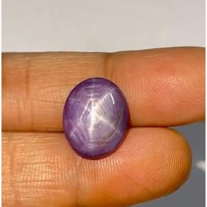 Puede incluir: Una piedra preciosa cabujón de forma ovalada con un tono púrpura intenso y un patrón de estrella de seis puntas claramente visible en su superficie lisa y pulida. La piedra exhibe un efecto de chatoyance que crea un reflejo estelar.