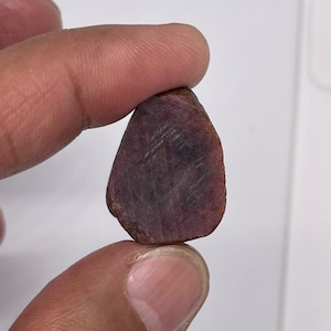 Op de afbeelding: Een ruwe, ongeslepen robijn met een diepe roodpaarse tint en een natuurlijke, onregelmatige vorm. De steen vertoont zichtbare interne insluitingen en een mat, licht getextureerd oppervlak, wat het organische en onbewerkte uiterlijk benadrukt.