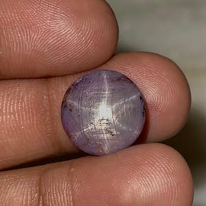 May include: A round, cabochon-cut star sapphire gemstone with a translucent, light purple hue. The stone displays a distinct six-rayed star effect, known as asterism, centred on its surface, with visible internal inclusions and fine striations.