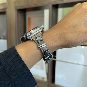 Puede incluir: Un reloj de pulsera con caja de metal plateado y correa de cuero marrón claro. La correa está decorada con dos tachuelas plateadas en forma de estrella y cuenta con dos trabillas de metal plateado para asegurar el extremo de la correa.