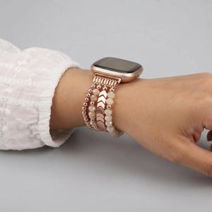 May include: A rose gold smartwatch band featuring multiple strands of small, round, light-colored beads and a central rose gold metal chevron-patterned link. The band is attached to a rectangular smartwatch with a dark, blank screen display.
