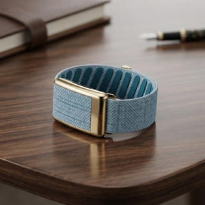 May include: A wearable fitness tracker featuring a light blue fabric band with a subtle grid pattern. The device includes a polished gold-tone metal clasp and housing, with a teal interior lining that has a series of vertical grip ridges.