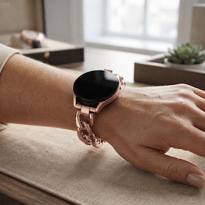 Puede incluir: Un reloj inteligente con pantalla redonda negra y correa de eslabones metálicos en oro rosa. El reloj presenta un acabado pulido en la caja y en los eslabones entrelazados de la pulsera, creando un aspecto coherente en la muñeca.