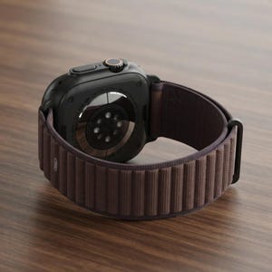 Puede incluir: Un reloj inteligente negro con un sensor circular en la parte posterior y una correa de tela acanalada de color marrón oscuro. La caja tiene un botón lateral y una corona giratoria con un detalle oscuro, con texto alrededor del sensor.