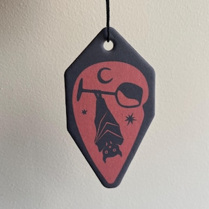 Op de afbeelding: Een ruitvormig hangend ornament met een zwarte rand en een rood midden. Het ontwerp toont het zwarte silhouet van een vleermuis die ondersteboven aan een wijnglassteel hangt, vergezeld door een maansikkel en twee kleine sterpictogrammen.