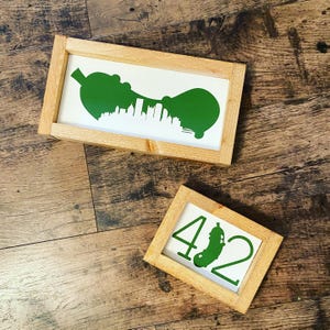 May include: Two framed art pieces with a wooden frame. One features a green pickle silhouette with a city skyline, the other displays the numbers 4 and 2 with a pickle graphic. Both are on a wooden surface.