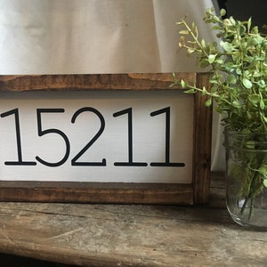 May include: A rustic wooden sign with the number "15211" in black against a white background. The sign is framed in dark brown wood. A glass jar with green foliage sits to the right.