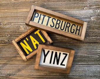 Op de afbeelding: Drie houten borden met zwarte letters op een houten achtergrond. De borden lezen "Pittsburgh", "N'at" en "Yinz".