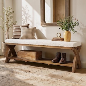 May include: A wooden bench with an X-shaped base and a white textured cushion seat. The bench features a lower storage shelf holding a rectangular wooden box and a pair of brown leather boots. Two decorative pillows sit on the bench seat.