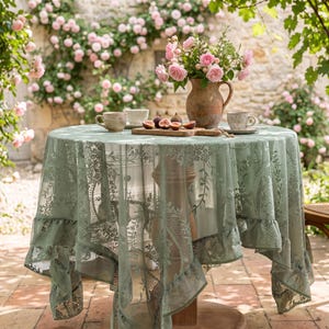 Könnte beinhalten: Ein runder Tisch mit einer salbeigrünen Spitzentischdecke mit Blumenmustern und einem gerüschten Rand. Auf dem Tisch stehen ein brauner Keramikkrug mit rosa Rosen, zwei Keramiktassen und ein Holzbrett mit aufgeschnittenen Feigen.