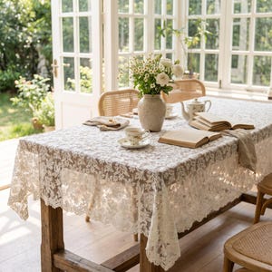 Pode incluir: Uma mesa de jantar retangular coberta com uma toalha de mesa de renda branca com um padrão floral intrincado. Sobre a mesa há um vaso de cerâmica cheio de margaridas brancas, uma xícara de chá, um bule e dois livros abertos.