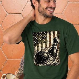 Może przedstawiać: Ciemnozielony t-shirt z krótkim rękawem z nadrukiem graficznym przedstawiającym czarną kulę do kręgli i pojedynczy kręgiel na tle postarzanego, zniszczonego wzoru flagi amerykańskiej z białymi gwiazdami i pasami na teksturowanym tle.
