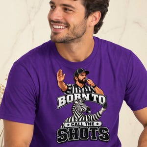 May include: A purple short-sleeve t-shirt featuring a graphic of a referee and a zebra, both wearing striped shirts. The referee holds a hand up and a whistle. The text reads BORN TO CALL THE SHOTS in bold white and black lettering.