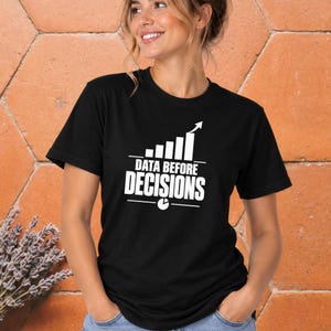 May include: A black short-sleeved t-shirt featuring a white graphic print on the chest. The design includes a bar chart with an upward arrow, the text "DATA BEFORE DECISIONS" in bold capital letters, and a small pie chart icon below the text.