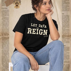 Pode incluir: Uma t-shirt preta de manga curta com o texto "LET DATA REIGN" numa fonte desgastada de cor branco sujo, com um pequeno ícone de coroa acima da palavra "REIGN" e uma pequena caixa "GS VERIFIED" no canto inferior direito da estampa.