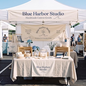 May include: A white pop-up canopy tent featuring a banner that reads Blue Harbor Studio Handmade Canvas Goods. The booth displays various canvas bags, small pouches, and folded textiles on a table covered with a beige cloth with the brand name.