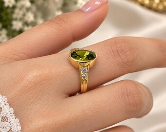 May include: A gold ring featuring a large, oval-cut olive green gemstone set in a smooth bezel. The band is crafted from polished gold and includes two small, clear, round-cut accent stones embedded vertically on each side of the central setting.
