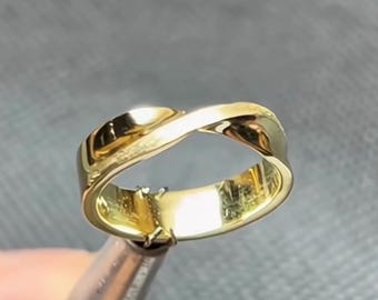 May include: A polished gold ring featuring a continuous twisted band design. The metal surface is smooth and reflective, creating a looped, infinity-like shape that curves around the finger opening. The band has a uniform width and thickness.