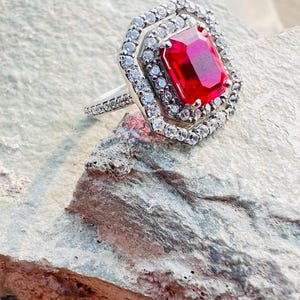 May include: A silver-toned ring featuring a large, rectangular, vibrant red center stone. The stone is surrounded by a double halo of small, clear, sparkling stones, with additional clear stones set along the visible portion of the ring band.