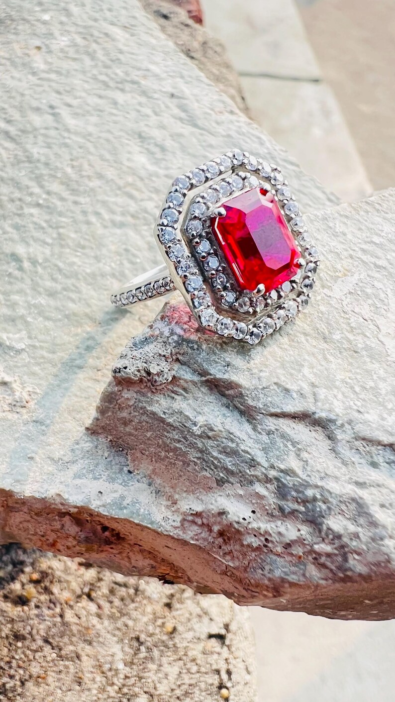 May include: A silver-toned ring featuring a large, rectangular, vibrant red center stone. The stone is surrounded by a double halo of small, clear, sparkling stones, with additional clear stones set along the visible portion of the ring band.