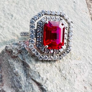 May include: A silver-toned ring featuring a large, rectangular, vibrant red center stone. The stone is surrounded by a double halo of small, clear, sparkling stones set in a geometric, octagonal frame. The band is thin and also set with small stones.