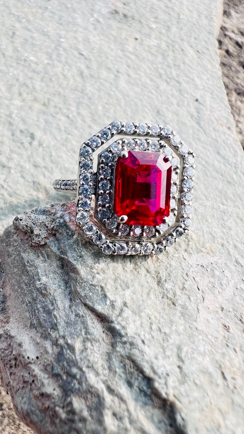 May include: A silver-toned ring featuring a large, rectangular, vibrant red center stone. The stone is surrounded by a double halo of small, clear, sparkling stones set in a geometric, octagonal frame. The band is thin and also set with small stones.