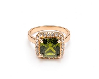 May include: A gold-toned ring featuring a square-cut olive green gemstone at the center. The stone is surrounded by a halo of small, clear, round-cut stones set in a square frame. The band is smooth, polished, and gold-toned metal throughout.