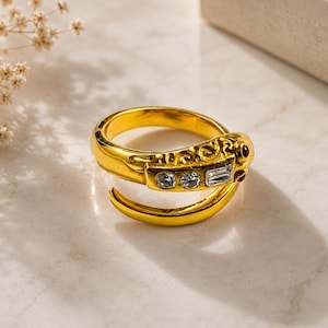 May include: A gold-toned wrap ring shaped like a snake with an engraved textured body. The head features two small round clear stones, one rectangular clear stone, and two tiny red stones for eyes, set against a polished metallic finish.
