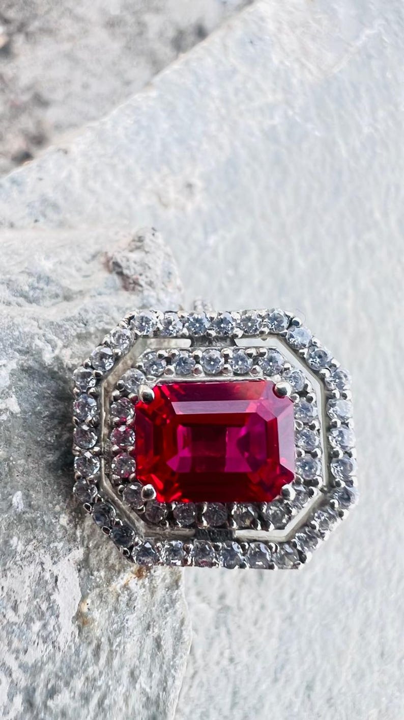 May include: A silver-toned ring featuring a large, rectangular, deep red center stone. The stone is surrounded by two concentric octagonal halos of small, clear, sparkling stones set in a metallic frame with a polished finish.