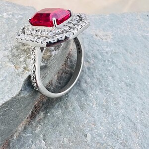 May include: A silver-toned ring featuring a large, rectangular, faceted red gemstone at the center. The stone is surrounded by a double halo of small, clear, round-cut stones, with additional clear stones set along the sides of the metal band.