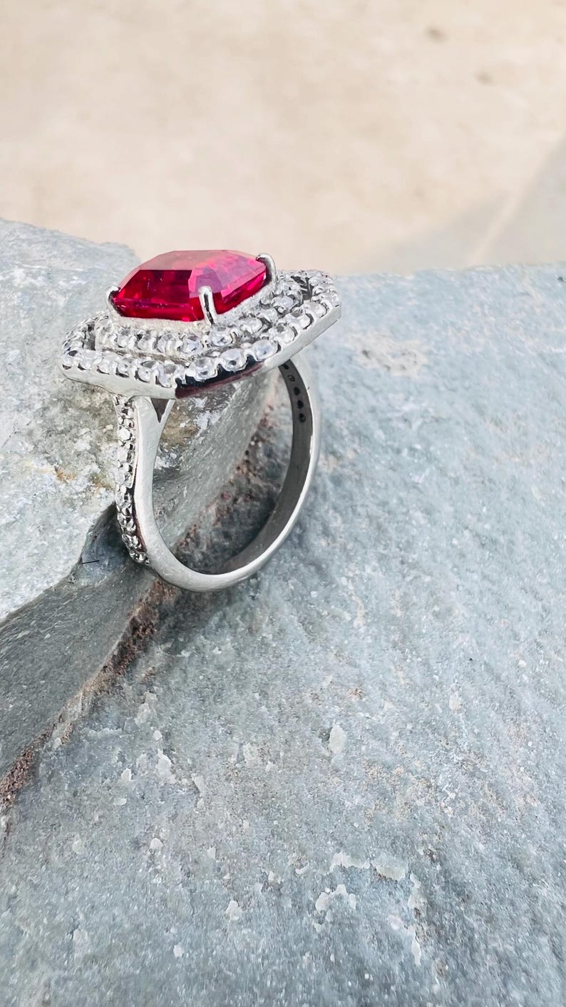 May include: A silver-toned ring featuring a large, rectangular, faceted red gemstone at the center. The stone is surrounded by a double halo of small, clear, round-cut stones, with additional clear stones set along the sides of the metal band.