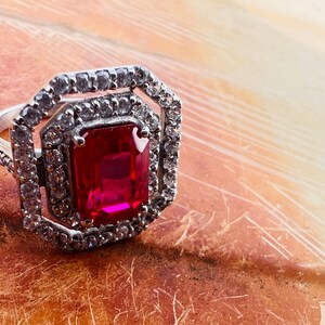 May include: A silver-toned ring featuring a large, rectangular, emerald-cut red center stone. The center stone is surrounded by two concentric octagonal halos of small, clear, sparkling stones, with additional clear stones set along the ring band.