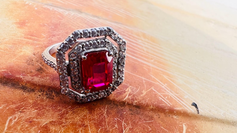 May include: A silver-toned ring featuring a large, rectangular, emerald-cut red center stone. The center stone is surrounded by two concentric octagonal halos of small, clear, sparkling stones, with additional clear stones set along the ring band.
