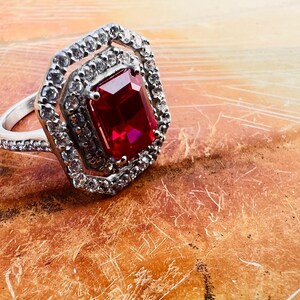 May include: A silver-toned ring featuring a large, rectangular, deep red center stone surrounded by a double halo of small, clear, sparkling stones. The band is also encrusted with small, clear stones, creating a bright, reflective appearance.
