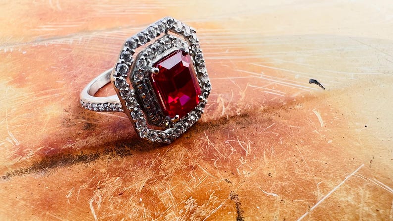May include: A silver-toned ring featuring a large, rectangular, deep red center stone surrounded by a double halo of small, clear, sparkling stones. The band is also encrusted with small, clear stones, creating a bright, reflective appearance.