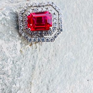 May include: A ring featuring a large, rectangular, vibrant red gemstone set in a silver-toned metal frame. The center stone is surrounded by two concentric octagonal halos of small, clear, sparkling white stones, creating a layered, geometric design.