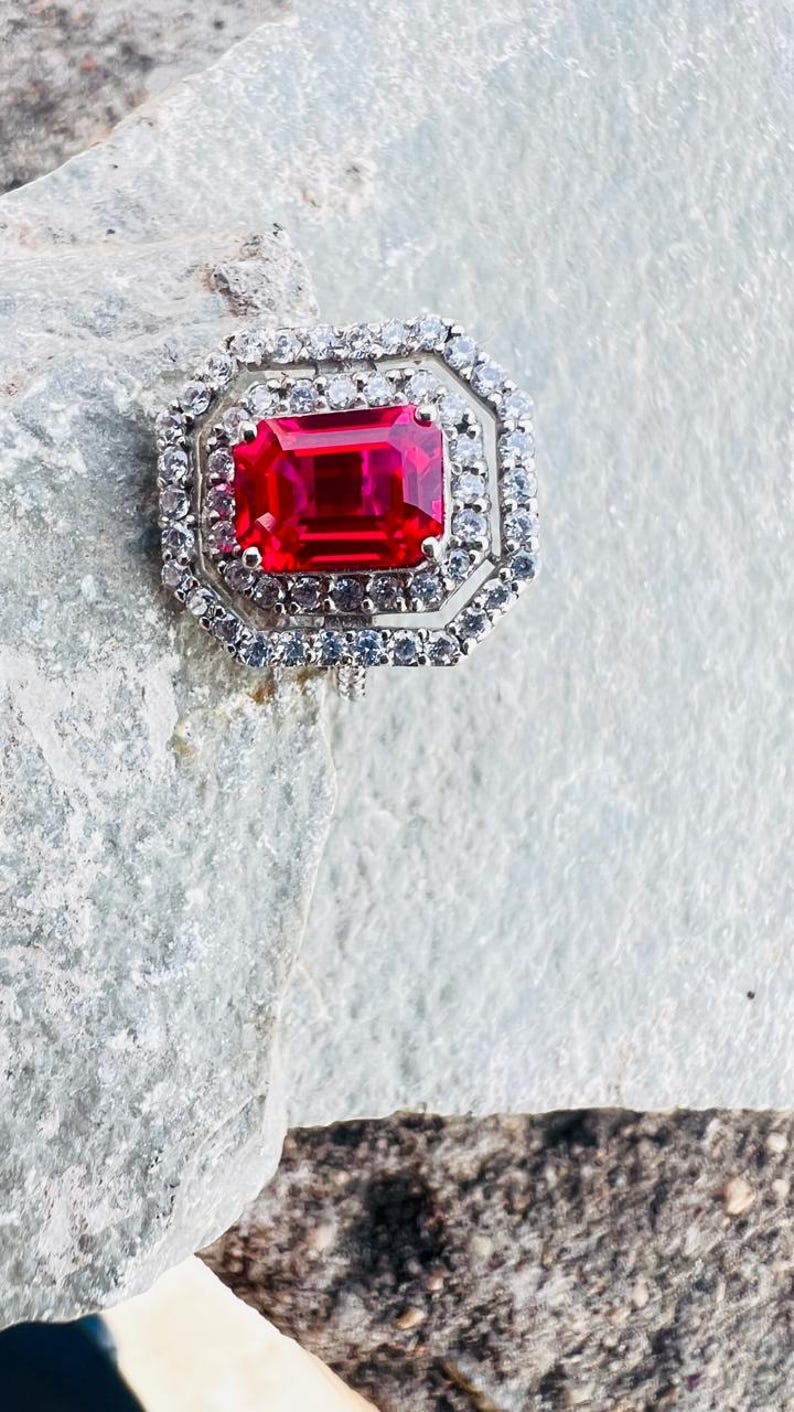 May include: A ring featuring a large, rectangular, vibrant red gemstone set in a silver-toned metal frame. The center stone is surrounded by two concentric octagonal halos of small, clear, sparkling white stones, creating a layered, geometric design.