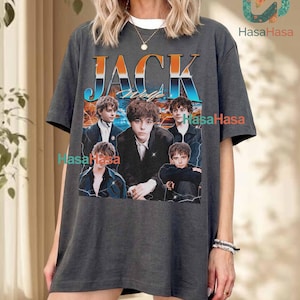 Puede incluir: Una camiseta gris carbón de manga corta con un estampado gráfico que incluye el texto JACK WOLF en letras con degradado naranja y azul. El diseño presenta un collage de cuatro fotografías de una persona joven con traje oscuro sobre un atardecer.