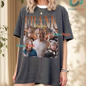 May include: A charcoal grey short-sleeve t-shirt featuring a graphic print of Frank Gallagher from the television show Shameless. The design includes the text FRANK GALLAGHER in a bold font above multiple images of the character in various outfits.