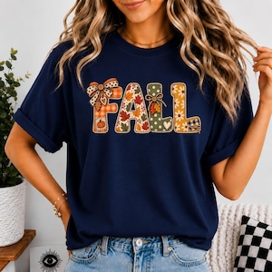 Könnte beinhalten: Ein dunkelblaues Kurzarm-T-Shirt mit dem Wort FALL in großen, dekorativen Buchstaben. Jeder Buchstabe hat ein einzigartiges Muster, darunter orangefarbenes Karomuster, Herbstblätter, grüne Punkte und gelbe Blumen mit kleinen Schleifen.
