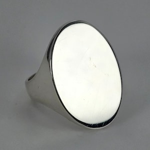 May include: A polished silver signet ring with a large, oval face. The ring has a smooth, reflective surface, and the band is thick and rounded. The ring is set against a neutral gray background.