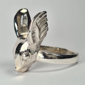 May include: A silver ring with a detailed rabbit head design. The rabbit's ears are pointed and have a textured surface. The ring is made of metal and has a smooth band.