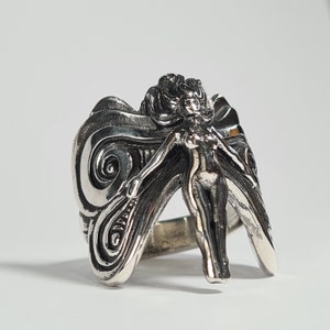 May include: A silver ring with a detailed design of a winged fairy. The fairy is depicted with flowing hair and a delicate body, and the wings are intricately sculpted with swirling patterns.